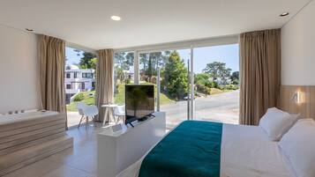 Una habitación de hotel moderna con una cama grande, un rincón de descanso con televisor y vistas a la calle y zonas verdes.