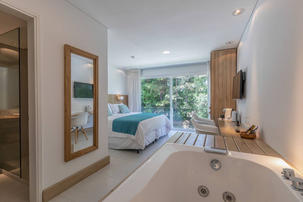 Een moderne hotelkamer met een groot bed, een zithoek met uitzicht, een badkuip en een balkon met uitzicht op de groene omgeving.