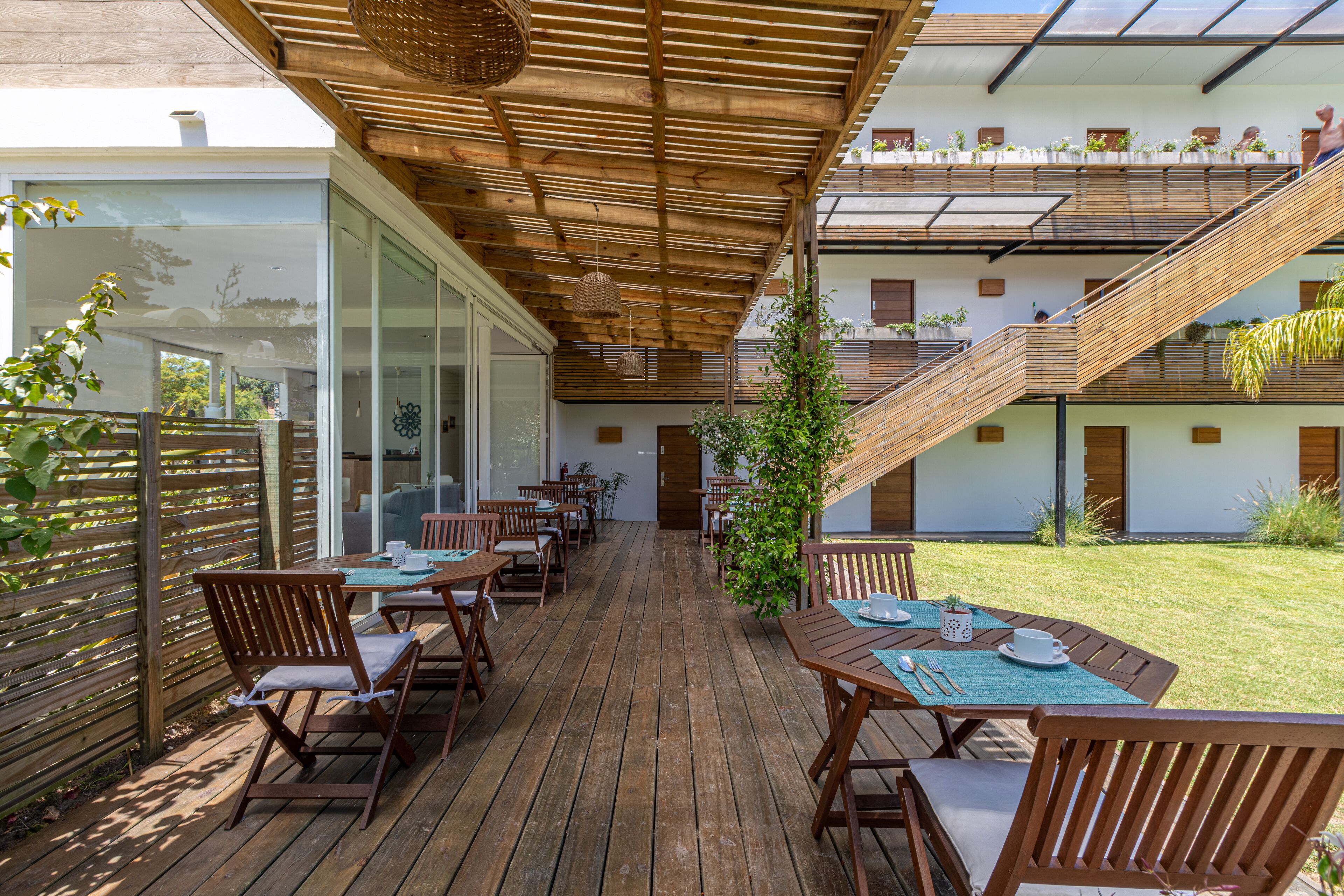 Una terraza de madera al aire libre con mesas y sillas, una escalera y vistas a un edificio con ventanas y plantas.