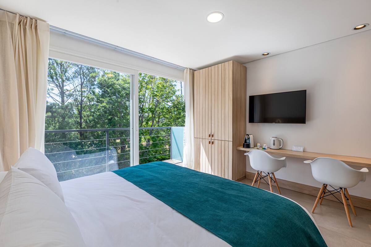Een moderne hotelkamer met een groot bed, een bureau met twee stoelen, een flatscreen televisie en een balkon met uitzicht op bomen.