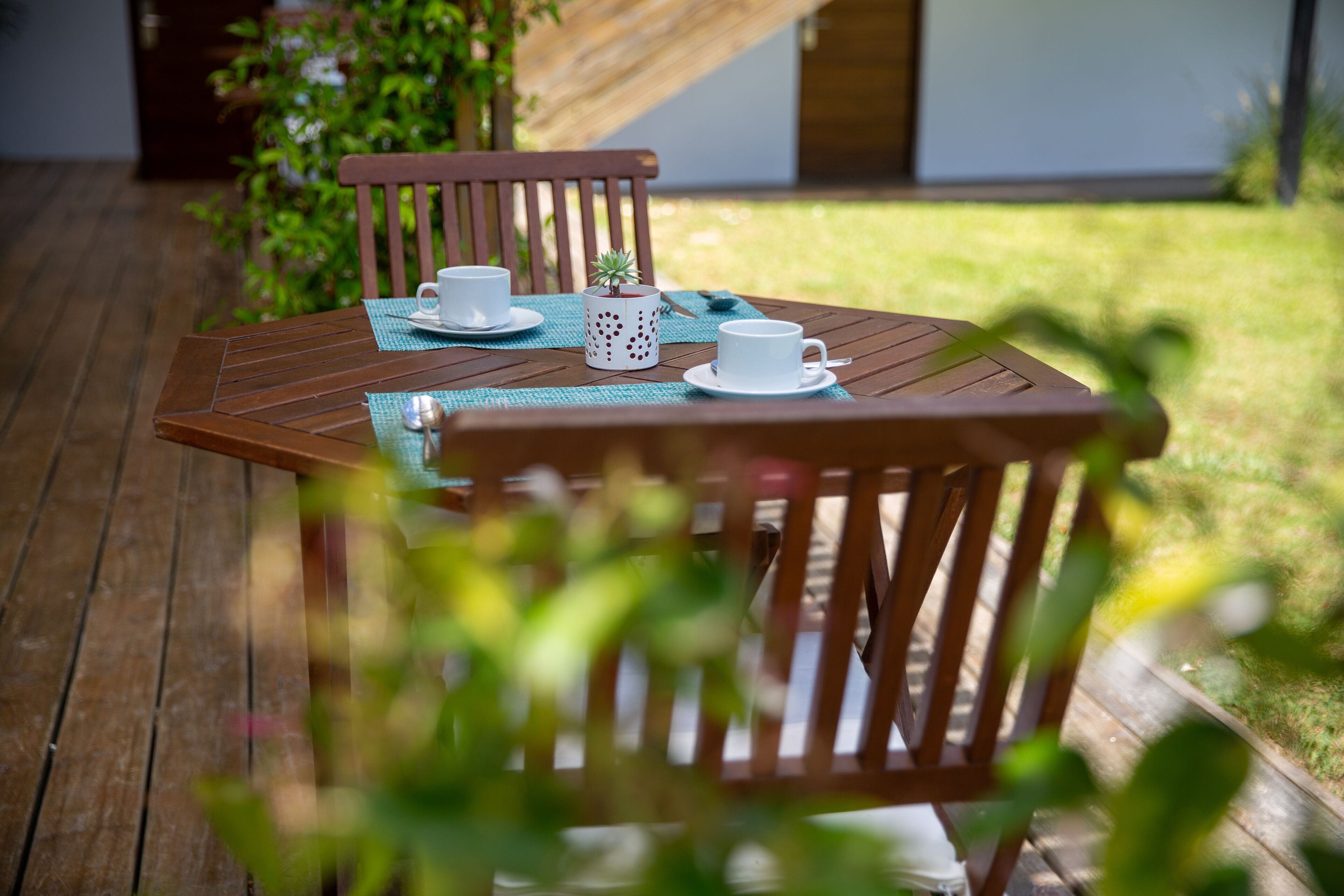 Una terraza de madera con una mesa dispuesta para dos personas, con dos tazas y platillos blancos, una planta decorativa y una taza de lunares.