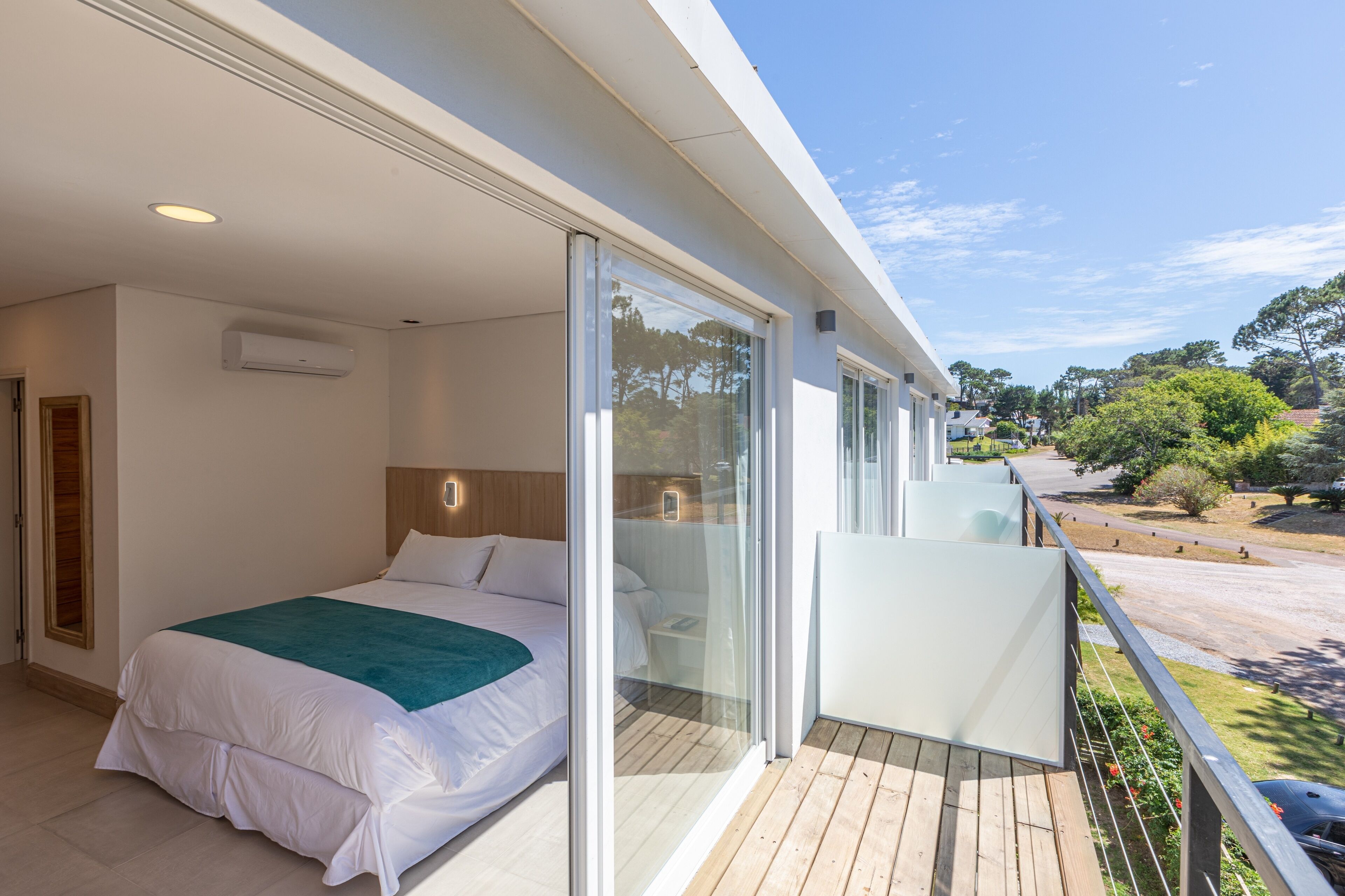 Een moderne slaapkamer met een groot bed, een balkon met een glazen deur en uitzicht op een parkeerplaats en bomen.