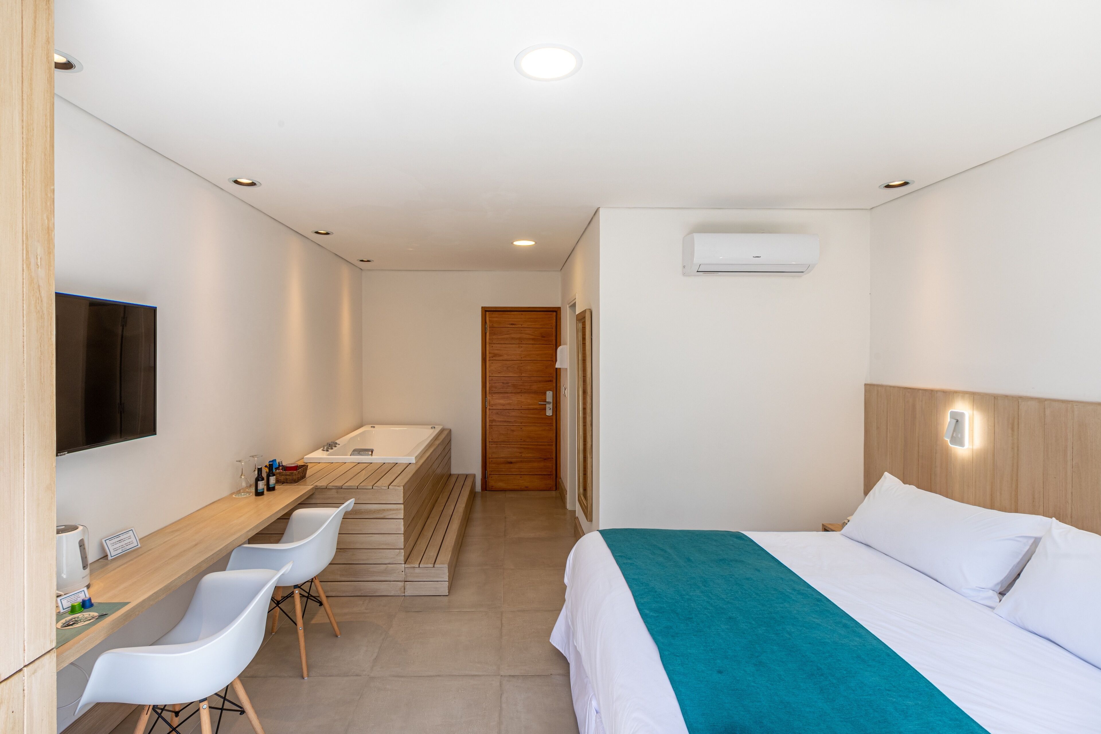 A hotel room with a bed, a wooden desk with chairs, a TV, a bathroom with a shower, and an air conditioning unit.