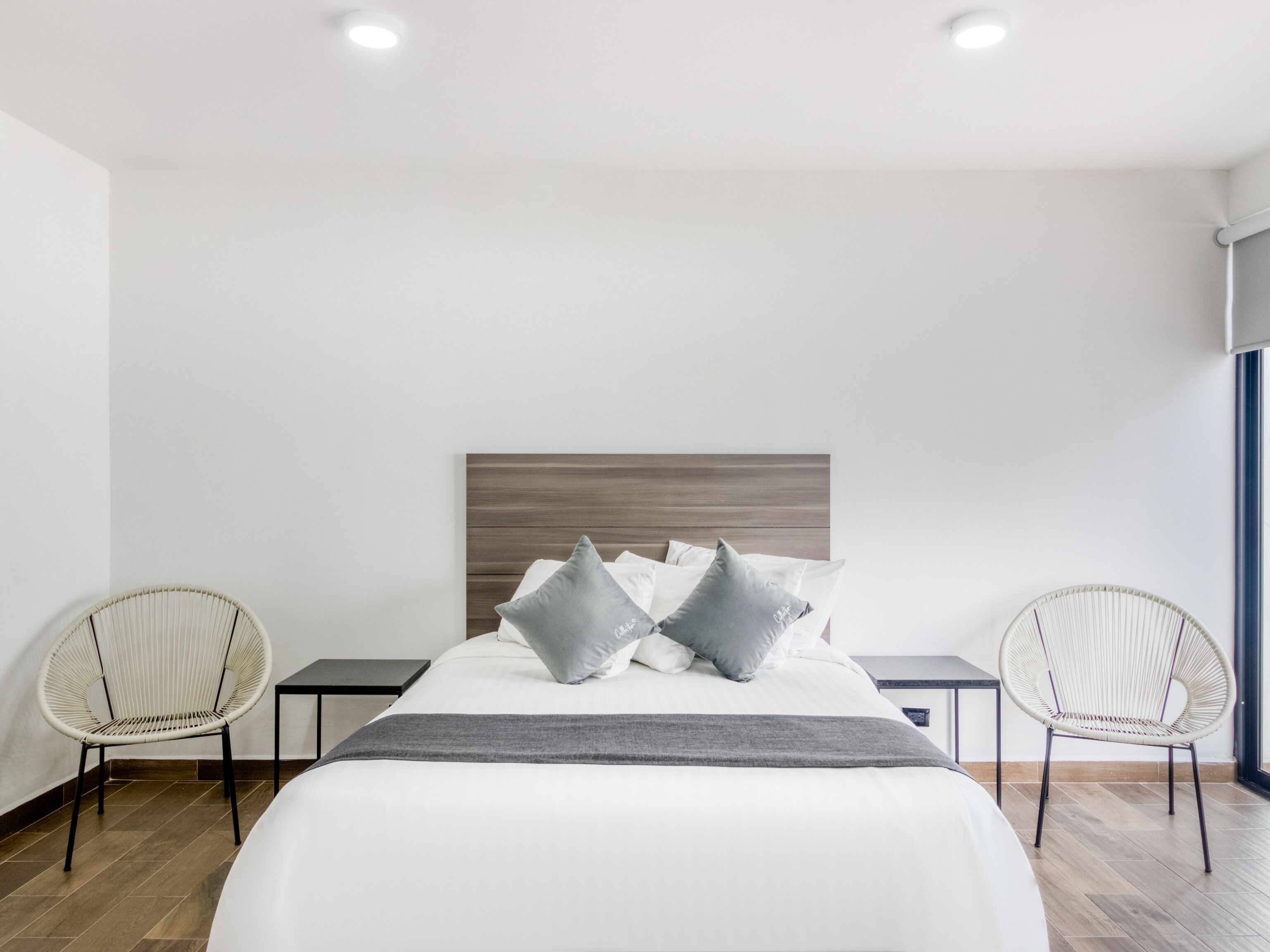 Quarto de hotel moderno com uma cama grande, duas cadeiras de vime, uma mesinha e vista para o exterior.