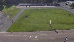 An aerial view of a large, open parking lot with a grassy area in the center, surrounded by trees and buildings.