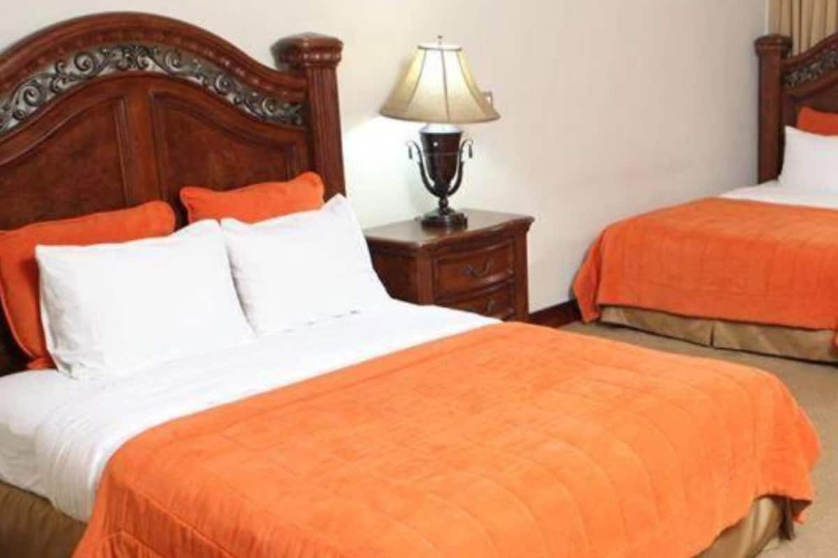 Ein Hotelzimmer mit zwei Betten, jeweils mit einer orangefarbenen Bettdecke, weißen Kissen und hölzernen Kopfteilen. Dazwischen befindet sich ein Nachttisch mit einer Lampe.