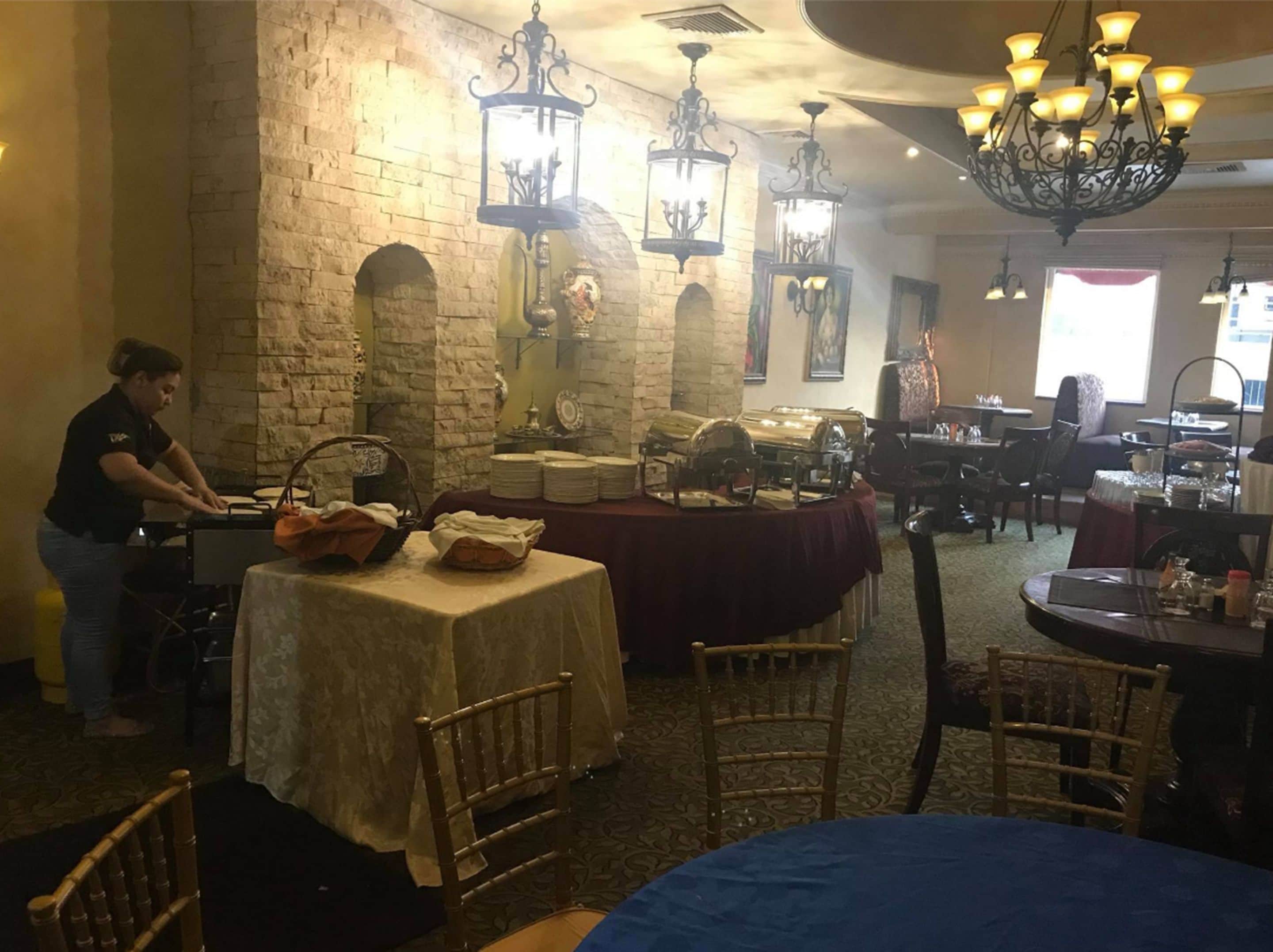 Un restaurante con paredes de piedra, faroles colgantes y una persona preparando comida. Las mesas están listas con platos y cestas.