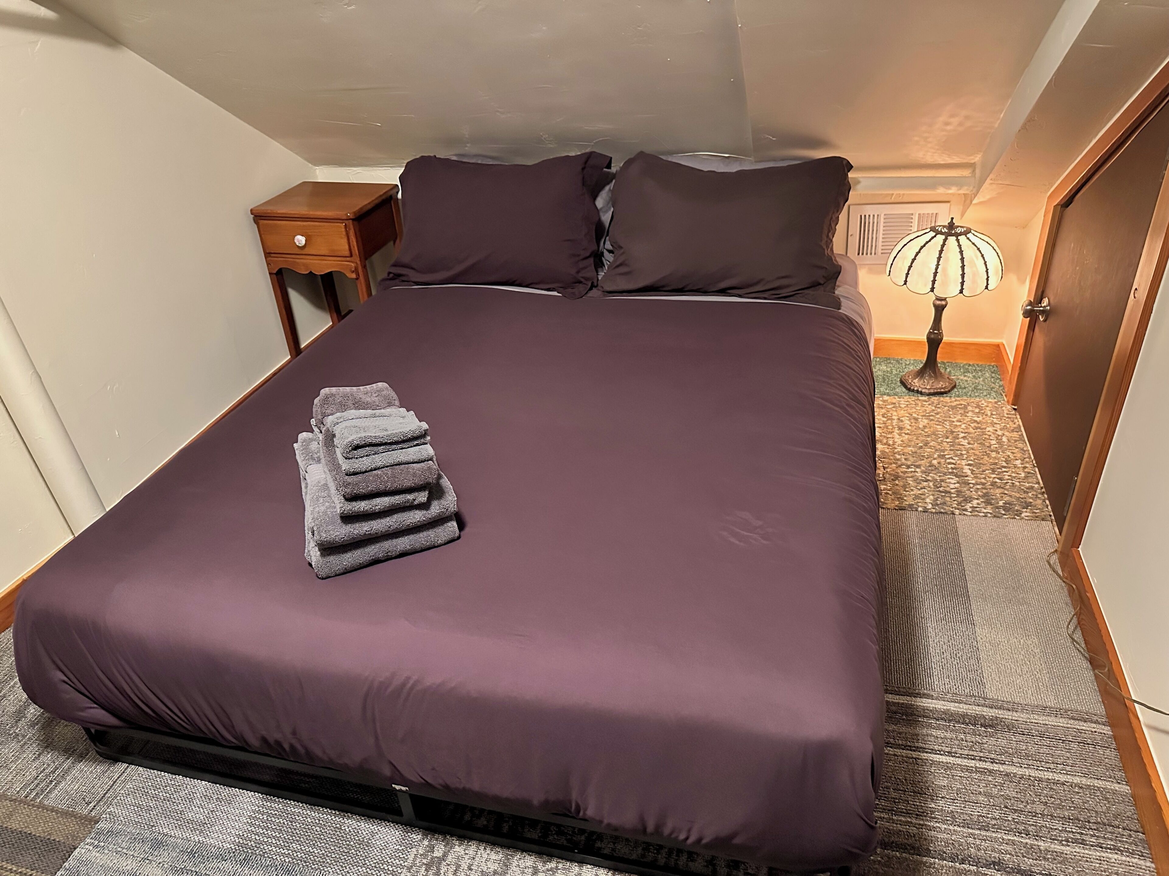 A neatly made bed with a purple bedspread and a stack of folded towels on the left side.