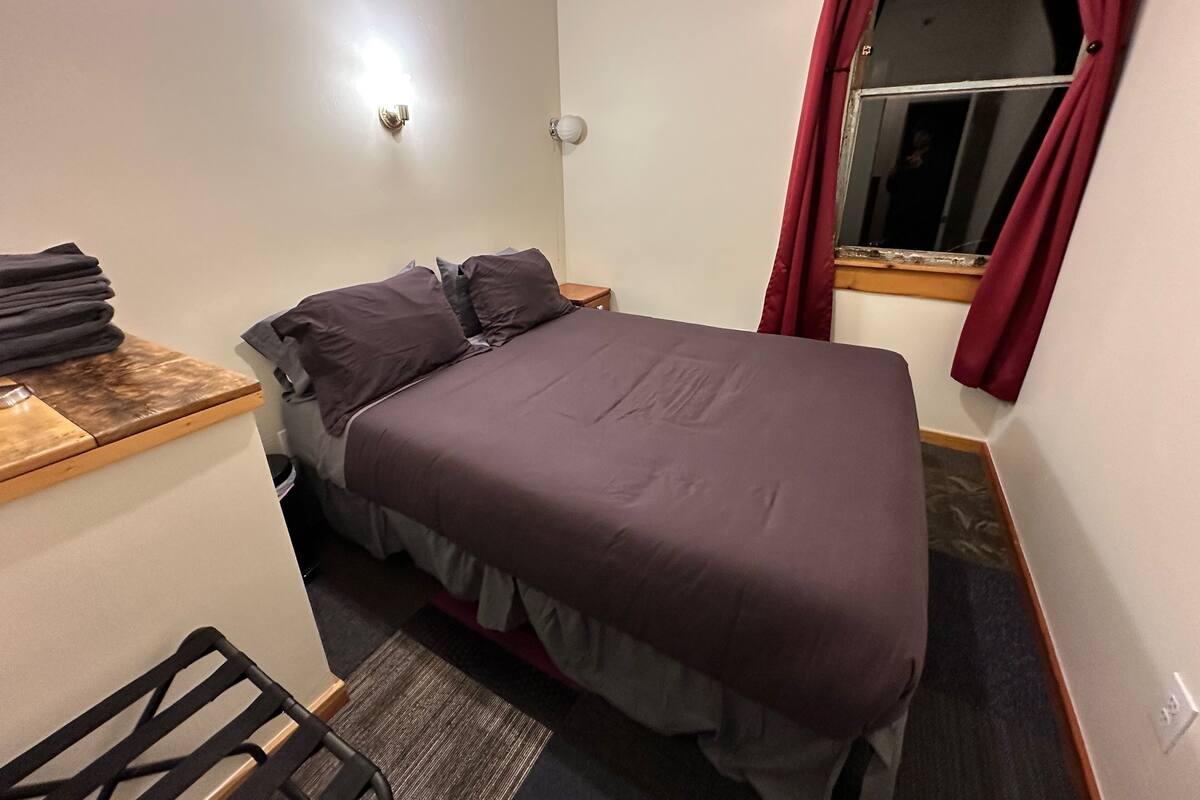 Une petite chambre à coucher, bien rangée, avec un lit, une petite table et une fenêtre avec des rideaux.