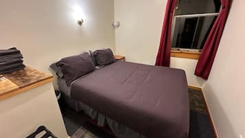 Um quarto pequeno e bem arrumado, com uma cama, uma mesinha e uma janela com cortinas.
