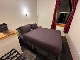 Um quarto pequeno e bem arrumado, com uma cama, uma mesinha e uma janela com cortinas.