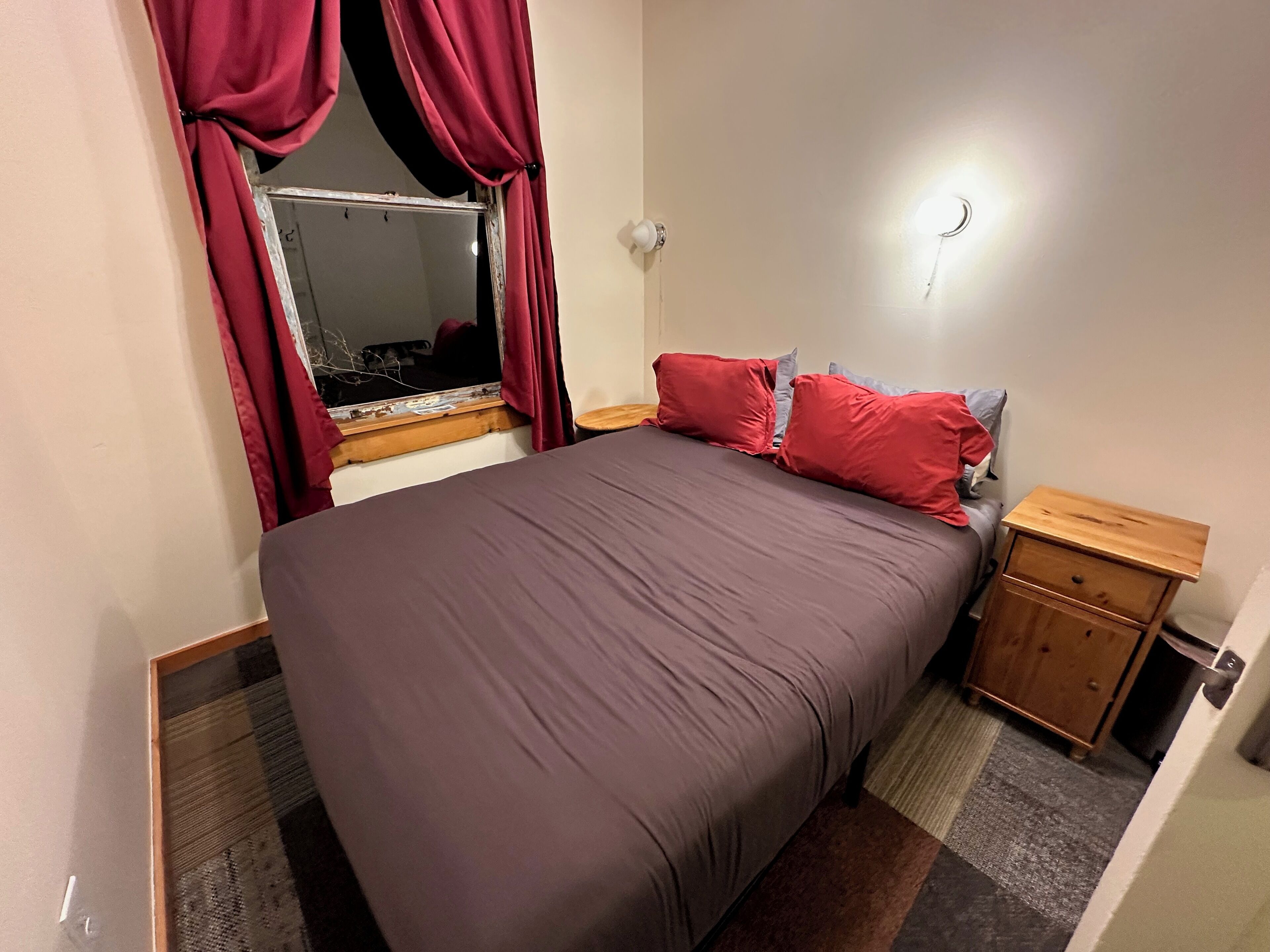 A bedroom with a large bed, red curtains, a wooden nightstand, and a small table.