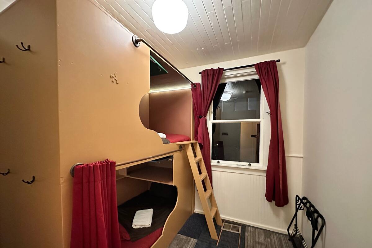 Une chambre avec un lit superposé, une échelle, une fenêtre avec des rideaux et une petite étagère.