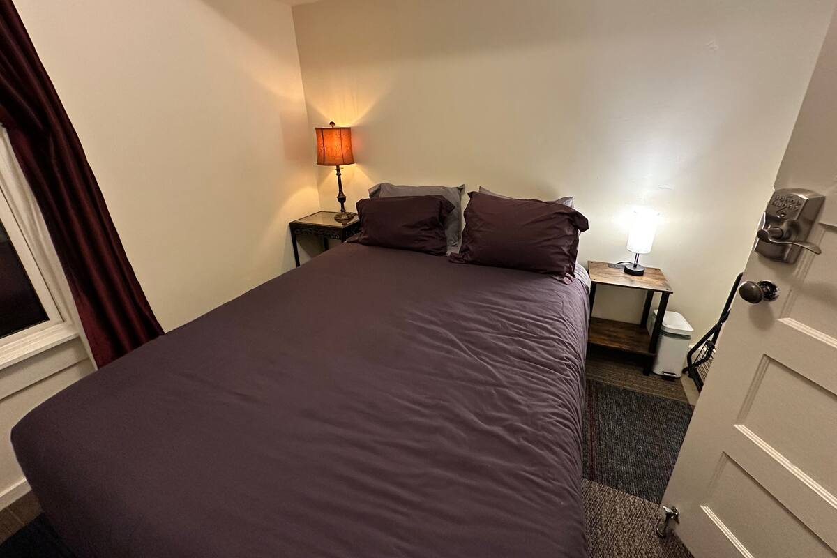 Une chambre d’hôtel avec un grand lit, une table de chevet avec une lampe, une porte dotée d’un système de verrouillage à clavier, et une fenêtre avec des rideaux.