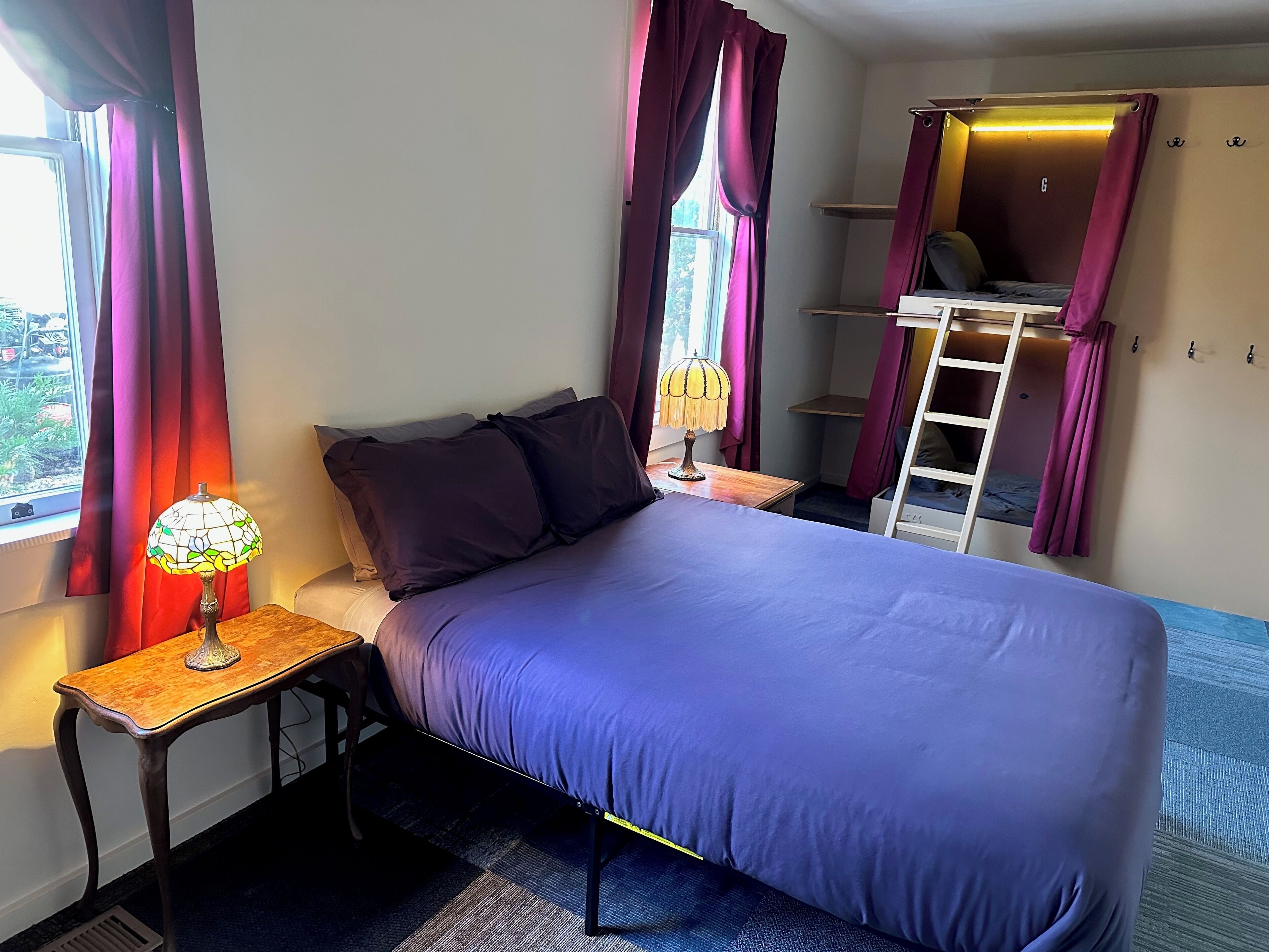 Une chambre à coucher comprenant un lit, une table de chevet, une lampe, une fenêtre avec des rideaux, une échelle et une étagère.