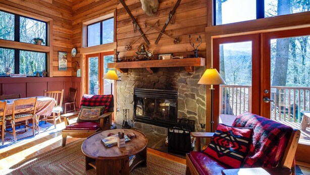 A cozy cabin interior with a stone fireplace, wooden furniture, and large windows offering a forest view. - 44mbr - Hot Tub- Pets Ok - Wifi - Bbq - Sleeps 8 2 Bedroom Home by RedAwning (Deming)