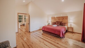 A bedroom with a wooden floor, a bed with a red bedspread, two bedside tables with lamps, and a door leading to another room. - 70mbr- Hot Tub - Bbq - Wi-fi - Pets Ok - Sleeps 8 3 Bedroom Home by RedAwning (Deming)