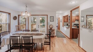 A dining area with a large table, chairs, and a chandelier. There is a wooden deck visible through the glass doors.