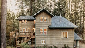 A two-story wooden cabin with a balcony, surrounded by dense forest.