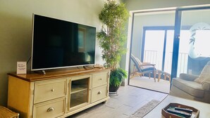A living room with a large flat-screen TV on a wooden cabinet, a potted plant, and a sliding glass door leading to an outdoor area with chairs.