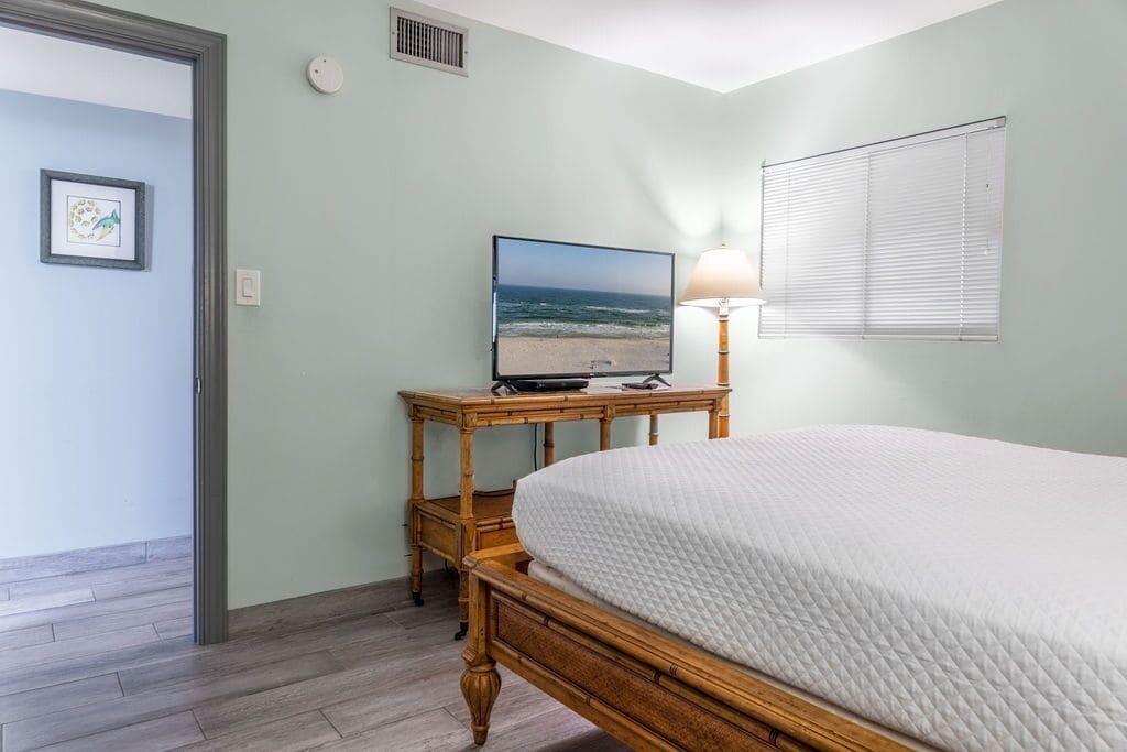 Ein Schlafzimmer mit einem Bett, einem Fernseher auf einem Holzständer, einer Lampe und einem Fenster mit Jalousien.