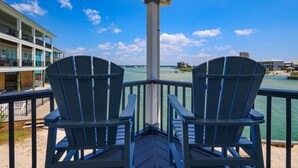 Deux fauteuils Adirondack sont placés sur une terrasse en bois, d’où l’on a une vue sur une étendue d’eau, avec des bâtiments visibles au loin.