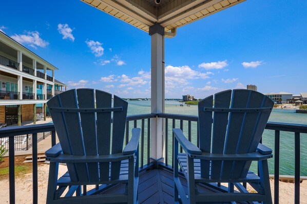 Two Adirondack chairs on a wooden deck overlooking a body of water with buildings in the background.