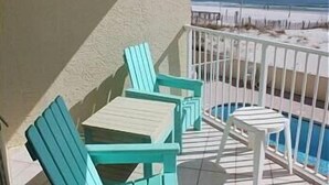 A balcony with turquoise chairs and a small table, overlooking a beach with a fence and a pool.