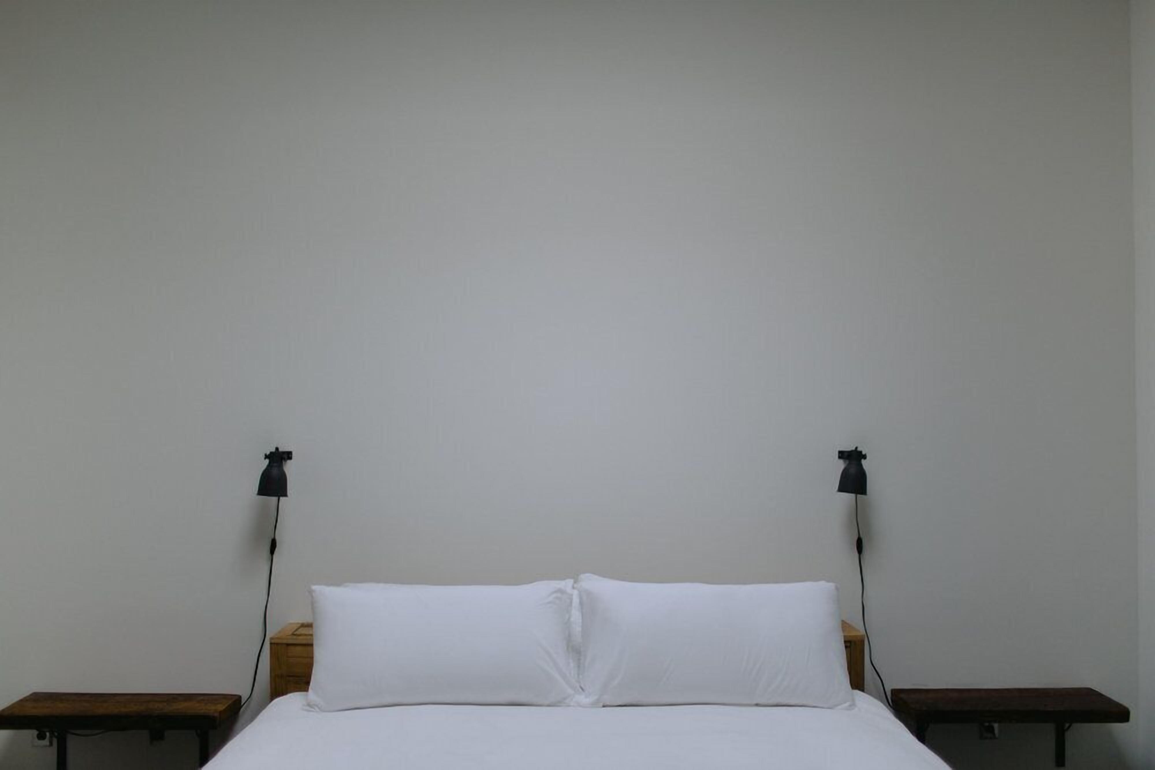 Une pièce simple et dépouillée, avec un lit simple, deux tables de chevet et deux lampes fixées au mur.