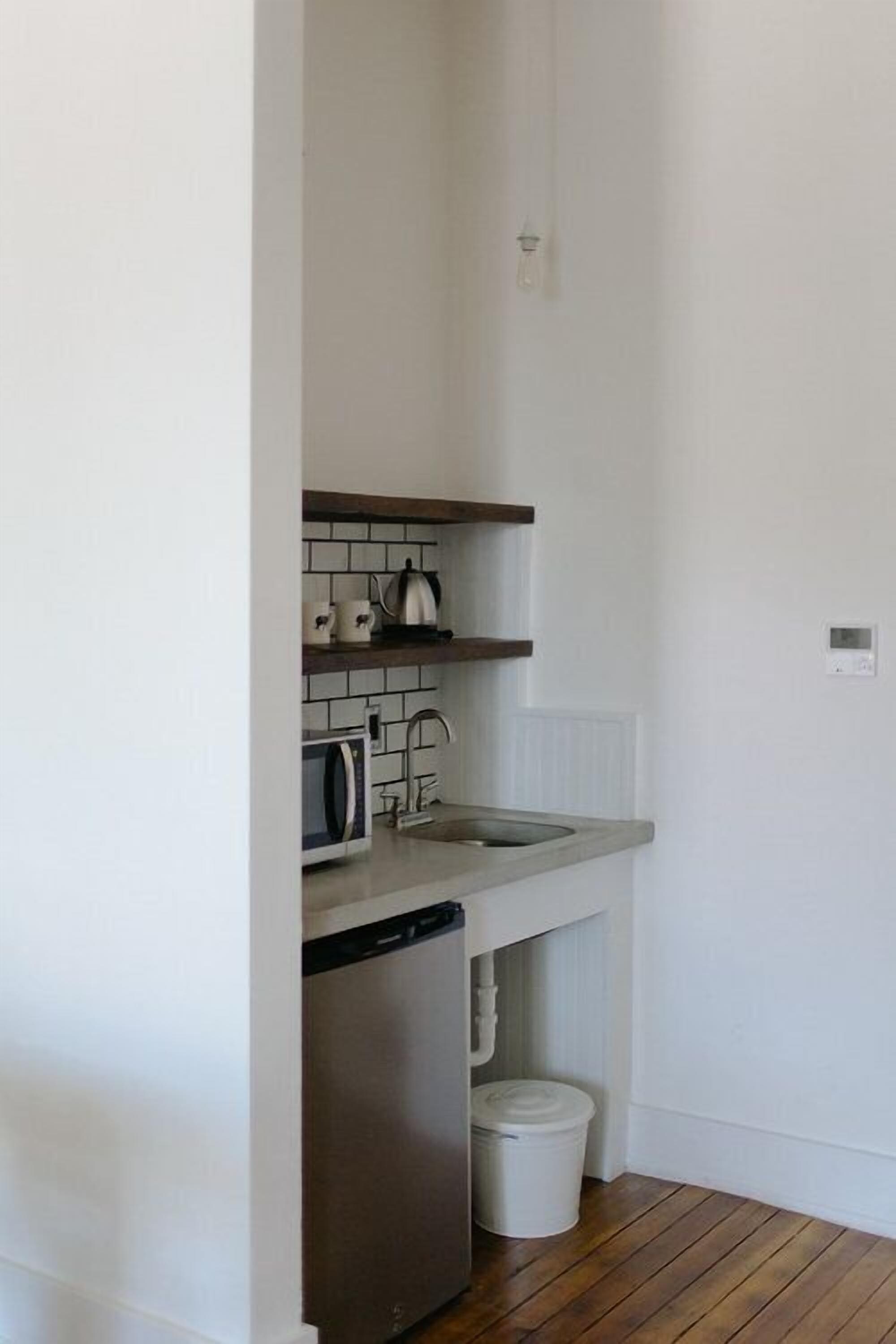 A compact kitchen with white walls, a wooden floor, a dishwasher, a sink, and a kettle on the countertop.