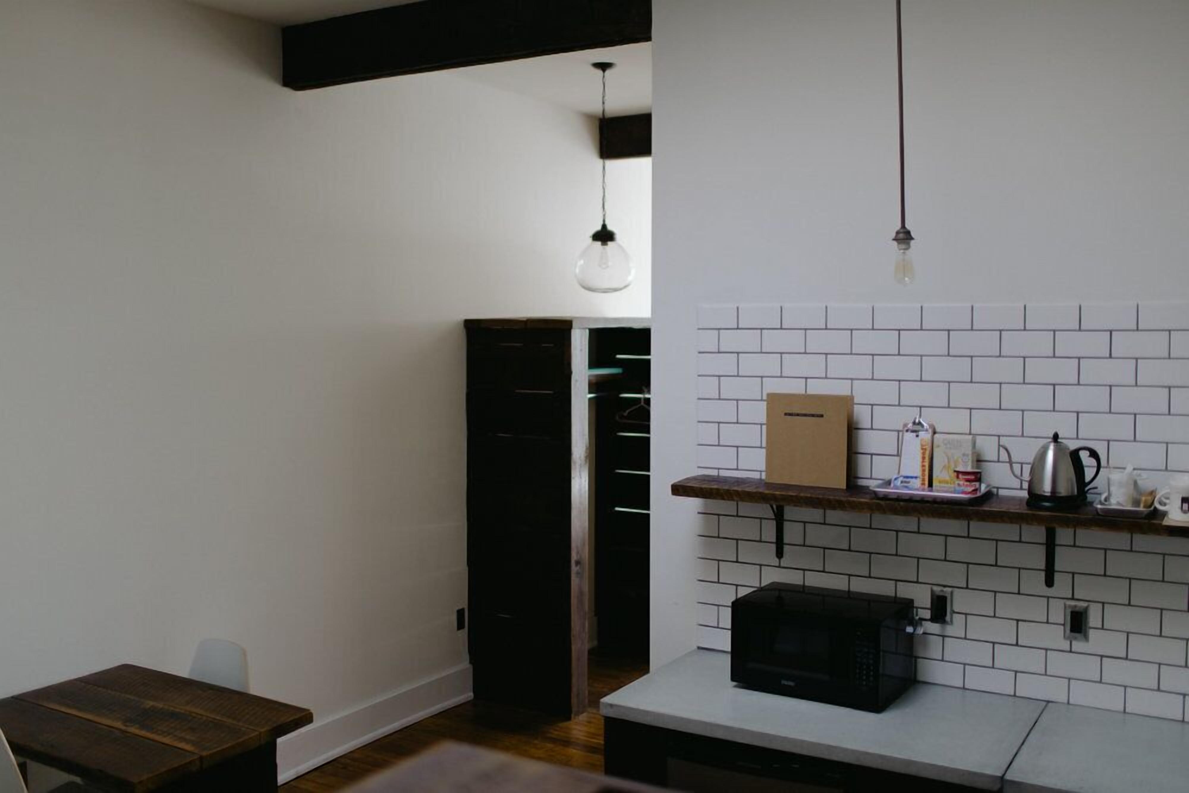 A compact kitchen with white tiled backsplash, a microwave, a wooden table, and a hanging light fixture.