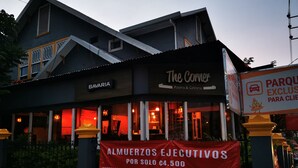 En bygning med skilt for 'The Corner Pizzeria & Cafeteria' og et banner, der reklamerer for 'Almuerzos Ejecutivos' for 4,50.