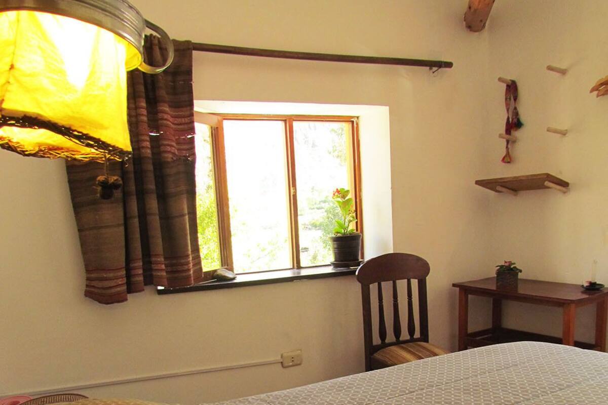 Un dormitorio con una cama, una silla, una mesita, una ventana con cortinas y una lámpara colgante.