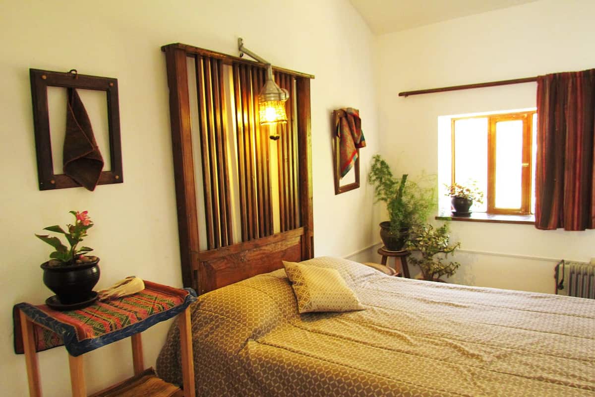 Un dormitorio con cabecera de madera, cama con ropa de cama estampada, una mesita con una planta y una ventana con cortinas.
