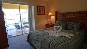 A hotel room with a bed, a nightstand, a lamp, a framed picture on the wall, and a view of an outdoor area with chairs and a table. - Beautiful Vacation Home Especially Convenient (Haines City)