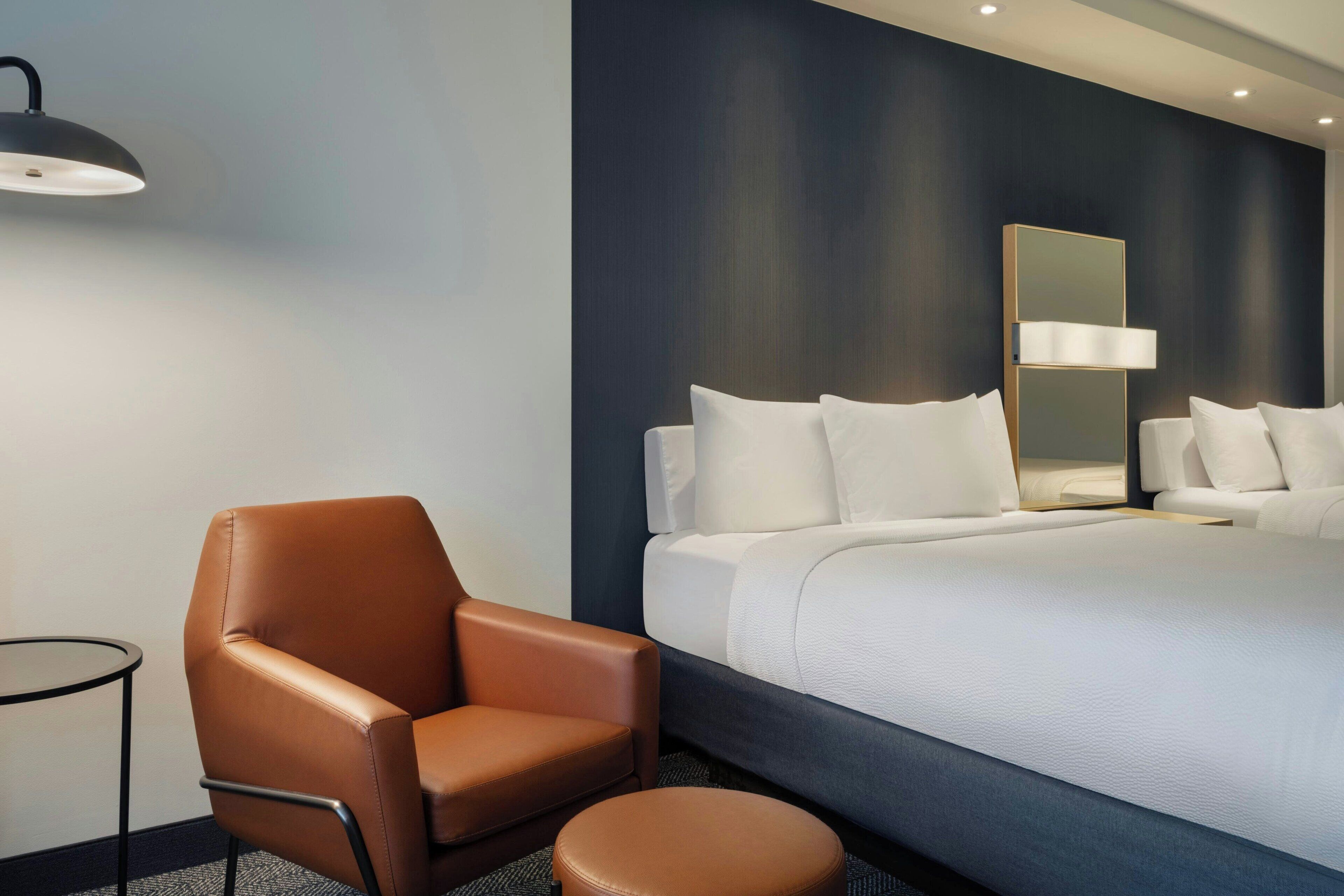 Habitación de hotel con una cama grande, un sillón de cuero marrón, una mesita redonda y una lámpara de pared.