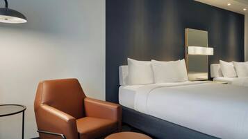 Habitación de hotel con una cama grande, un sillón de cuero marrón, una mesita redonda y una lámpara de pared.