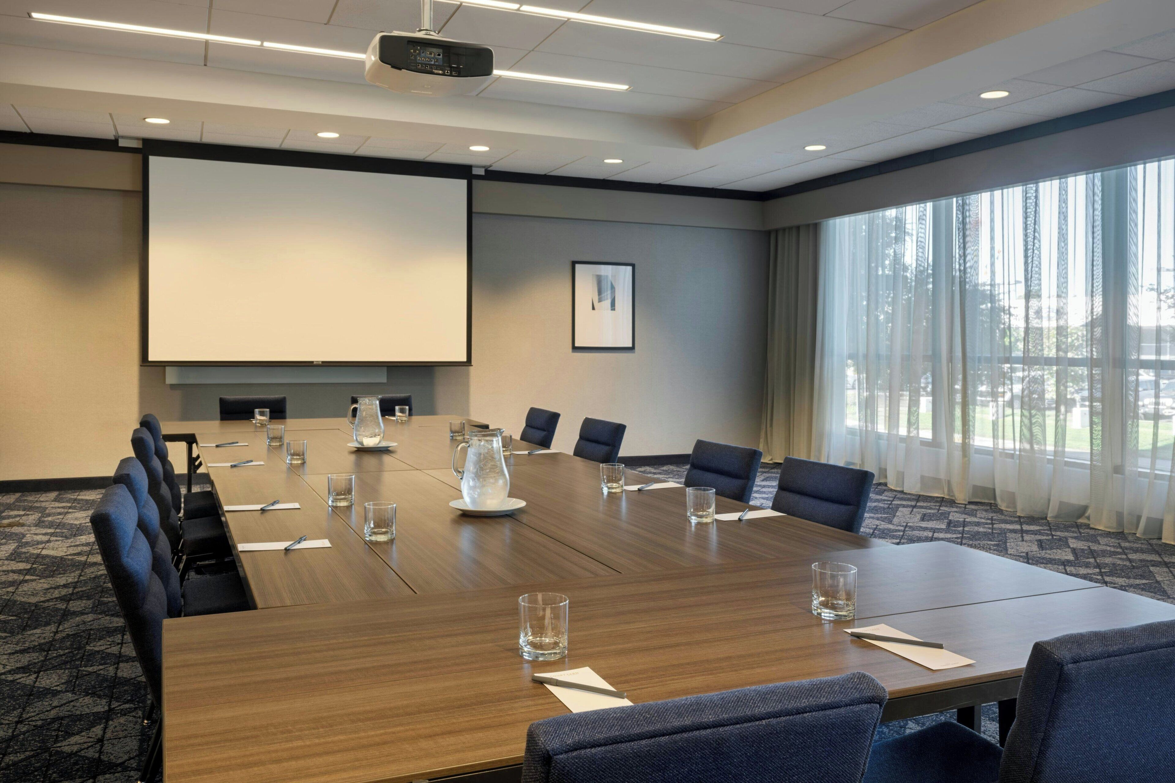 A conference room with a large table, chairs, a projector screen, and a large window with curtains.
