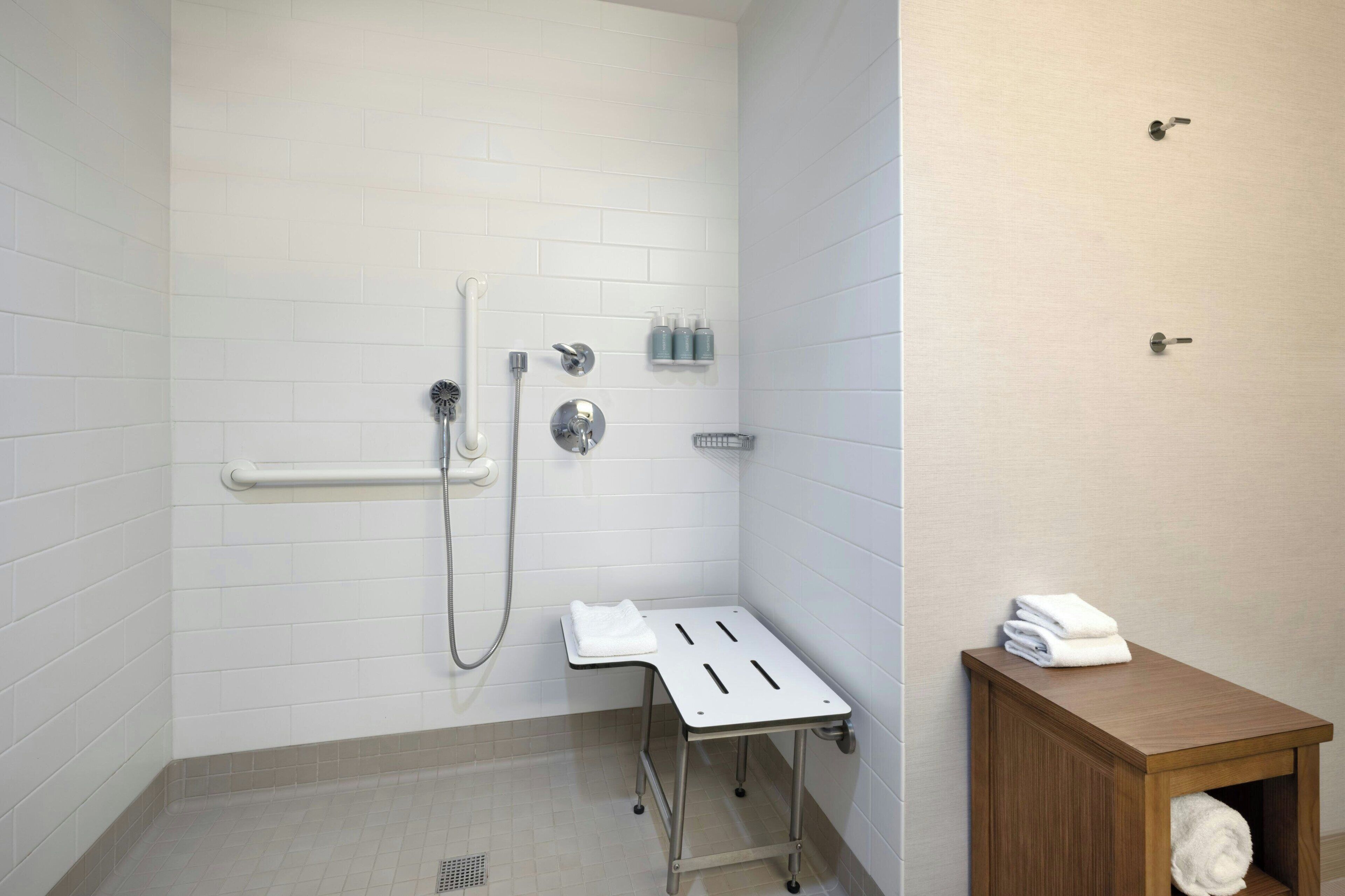 Un baño con paredes de azulejos blancos, una ducha con cabezal de mano, un perchero y un armario de madera con una toalla.