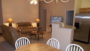 Un salon comprenant un canapé, deux fauteuils, une table basse et une table à manger avec quatre chaises. Un téléviseur est accroché au mur et un réfrigérateur se trouve au fond de la pièce.