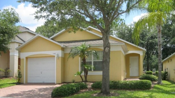 Une maison jaune à deux étages, avec une porte de garage blanche, un petit jardin devant la maison avec un arbre, et une pelouse bien entretenue.