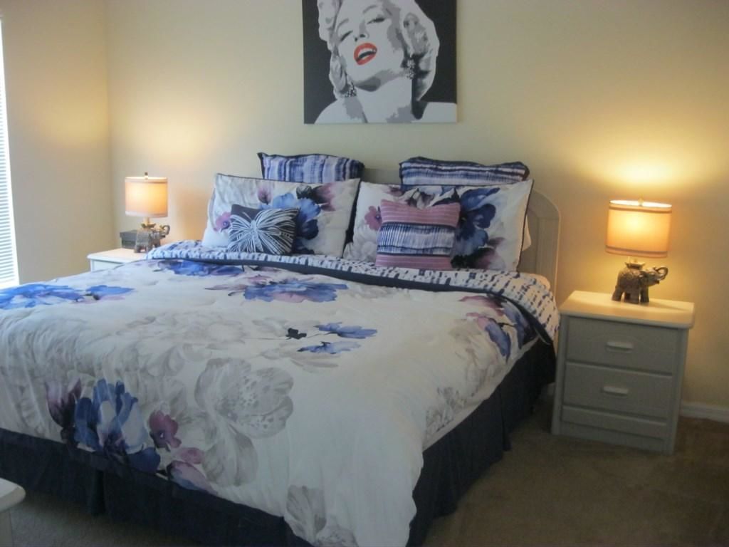 Een slaapkamer met een bloemenbedekking, twee bedlampjes, een nachtkastje met lamp, een lade en een poster aan de muur.