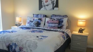 Une chambre à coucher avec une couvre-lit à motifs floraux, deux lampes de chevet, une table de chevet avec une lampe, un tiroir et une affiche accrochée au mur.