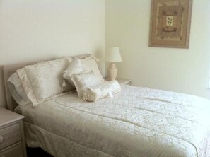 A neatly made bed with a white bedspread and pillows, a bedside table with a lamp, and a framed picture on the wall.