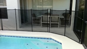 Une piscine entourée d’une clôture de sécurité noire, un espace patio avec une table et des chaises, et un coin salon couvert avec des coussins.