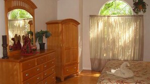 Ein Schlafzimmer mit einem Holzkoffer, einem Spiegel, einem Bett mit Schwanenkissen, einem Fenster mit durchsichtigen Vorhängen und einer Topfpflanze.