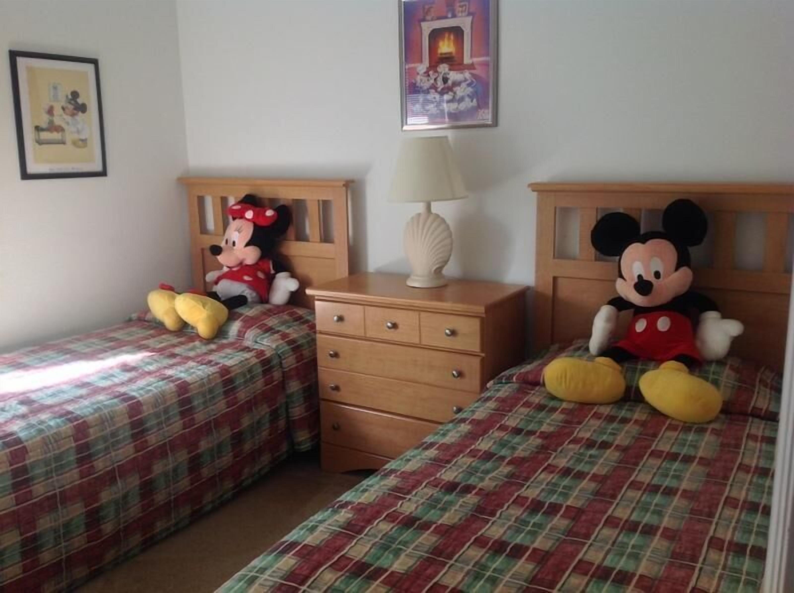 Une chambre avec deux lits, chacun orné d’une peluche à l’effigie de Mickey Mouse sur la tête de lit. Une table de chevet avec une lampe se trouve entre les deux lits.