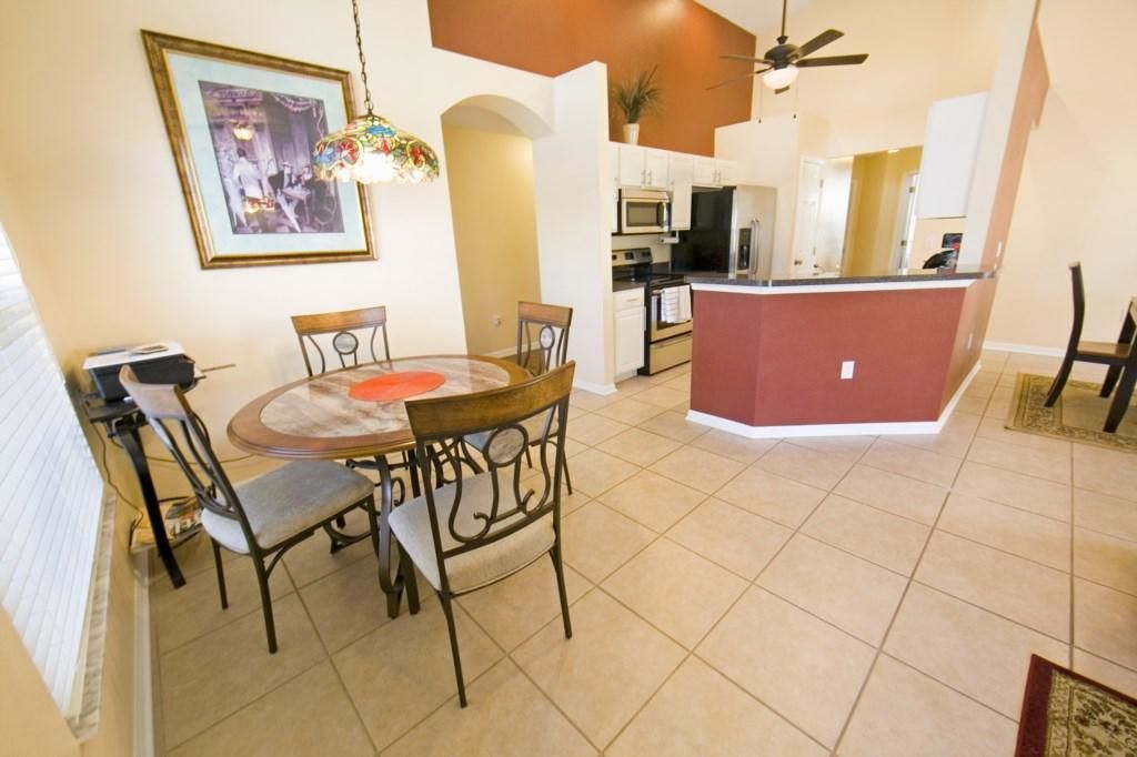 A dining area with a round table and chairs, a kitchen in the background, and a ceiling fan.
