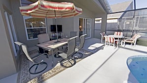 Une terrasse extérieure ensoleillée avec une piscine, une table entourée de chaises et un parasol à rayures.