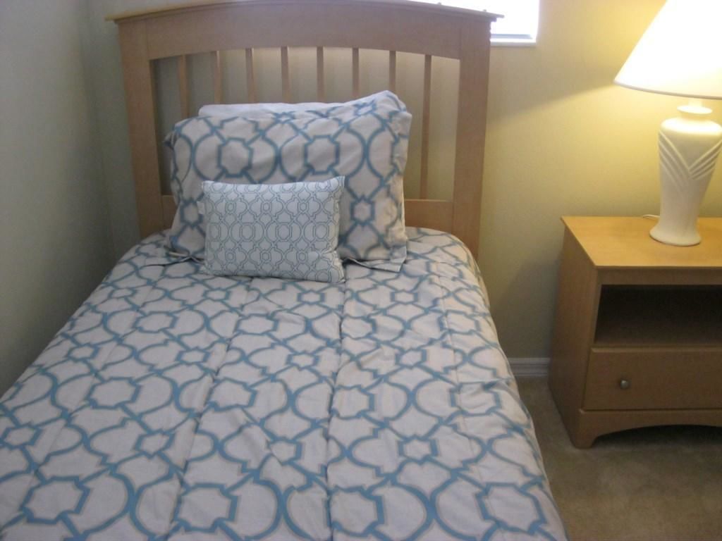 A bed with a patterned bedspread, a wooden headboard, a nightstand with a lamp, and a drawer.